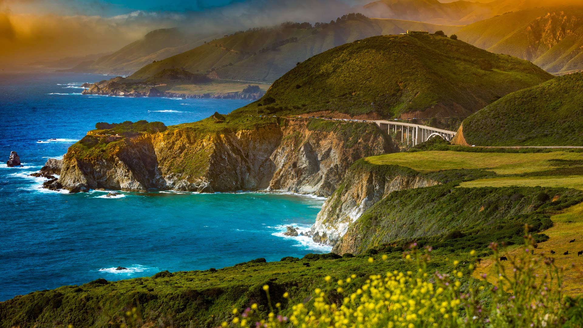 Landscape image of cliffs on the edge of the deep blue ocean with the sun setting and a bridge in the far distance.