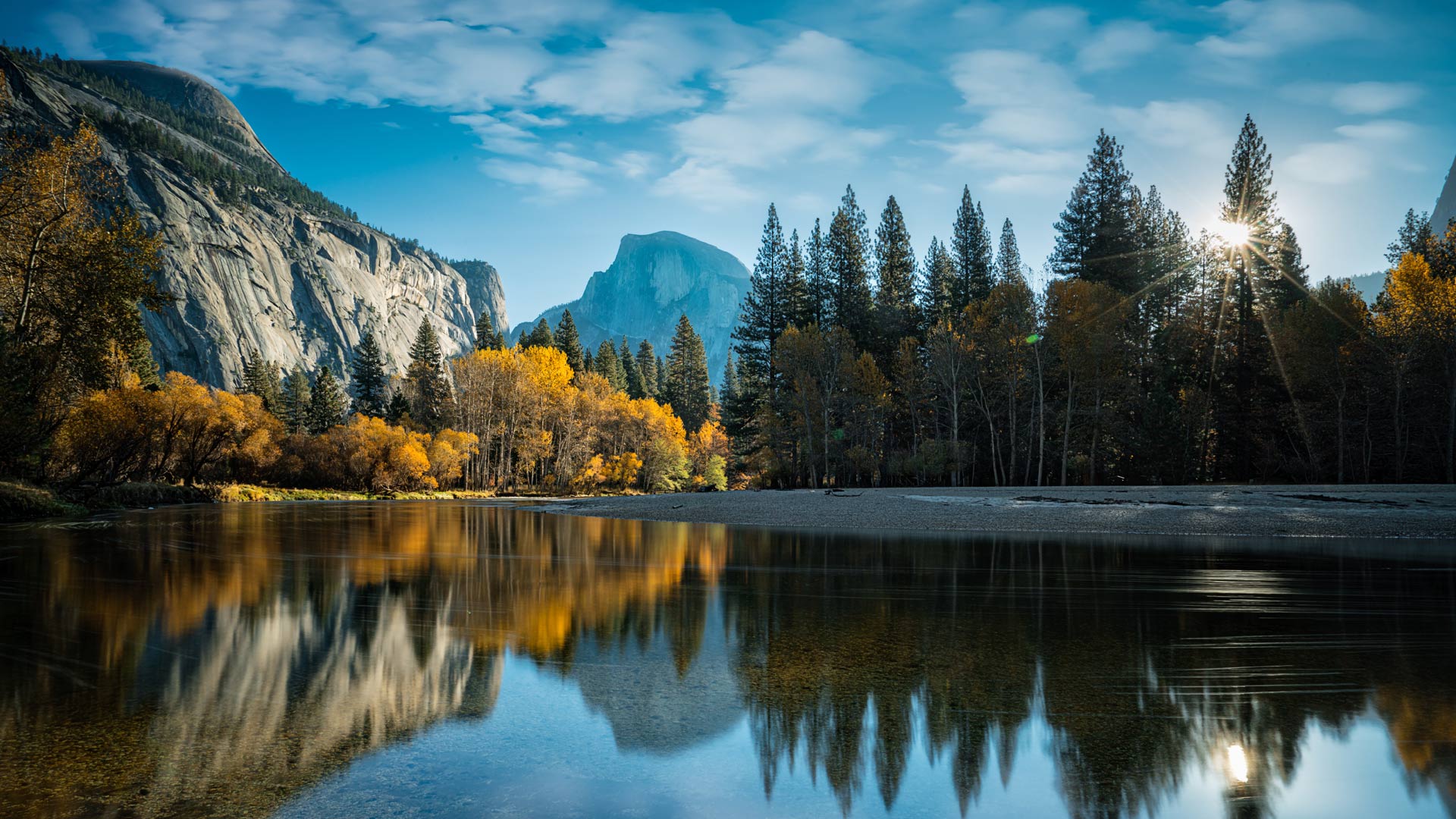 Landscape image of Yosemite Mountains in Autum reflecting on a body of water as sun peaks through the trees in a distance.